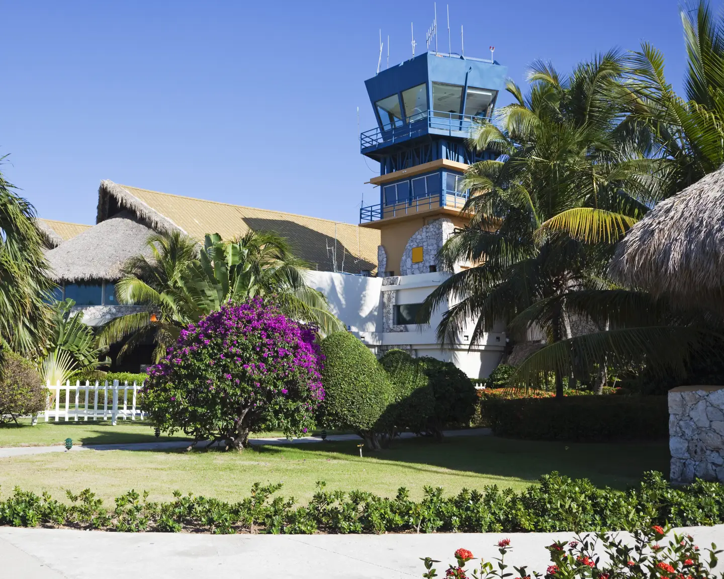 Un aeropuerto tropical con torre de control, techos de paja y plantas con flores en un ambiente caribeño.