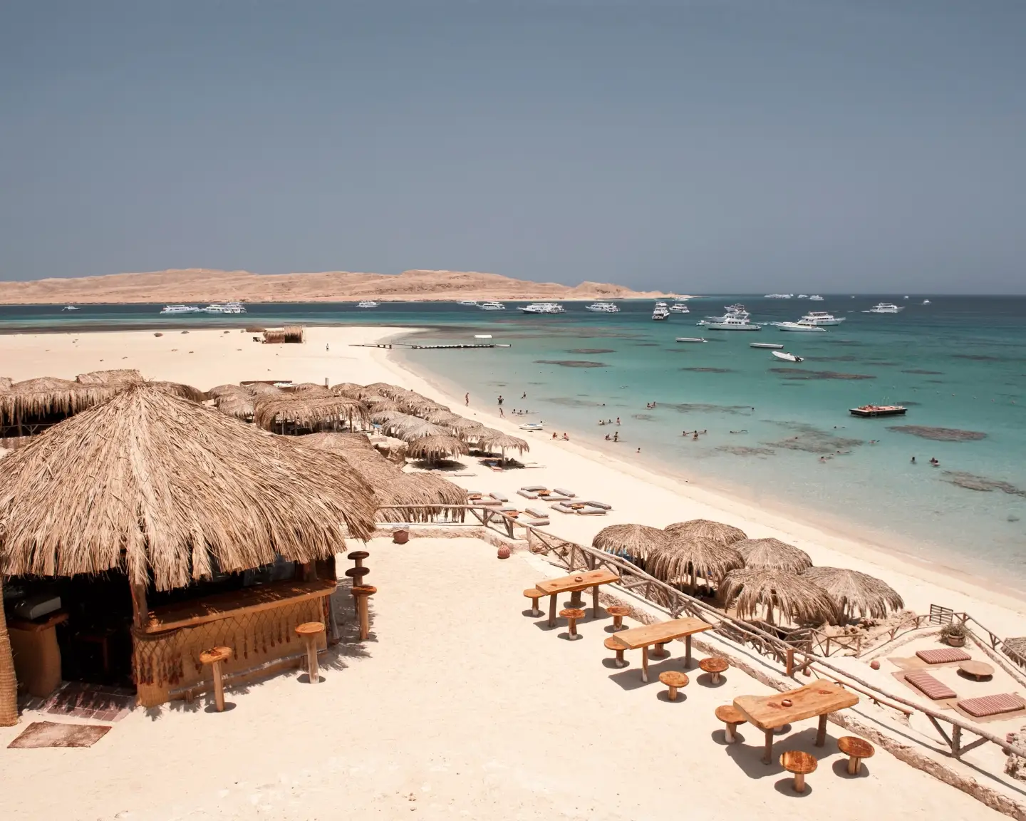 Sandstrand bei Hurghada mit strohgedeckten Strandhütten, türkisblauem Wasser und Ausflugsbooten im Roten Meer.
