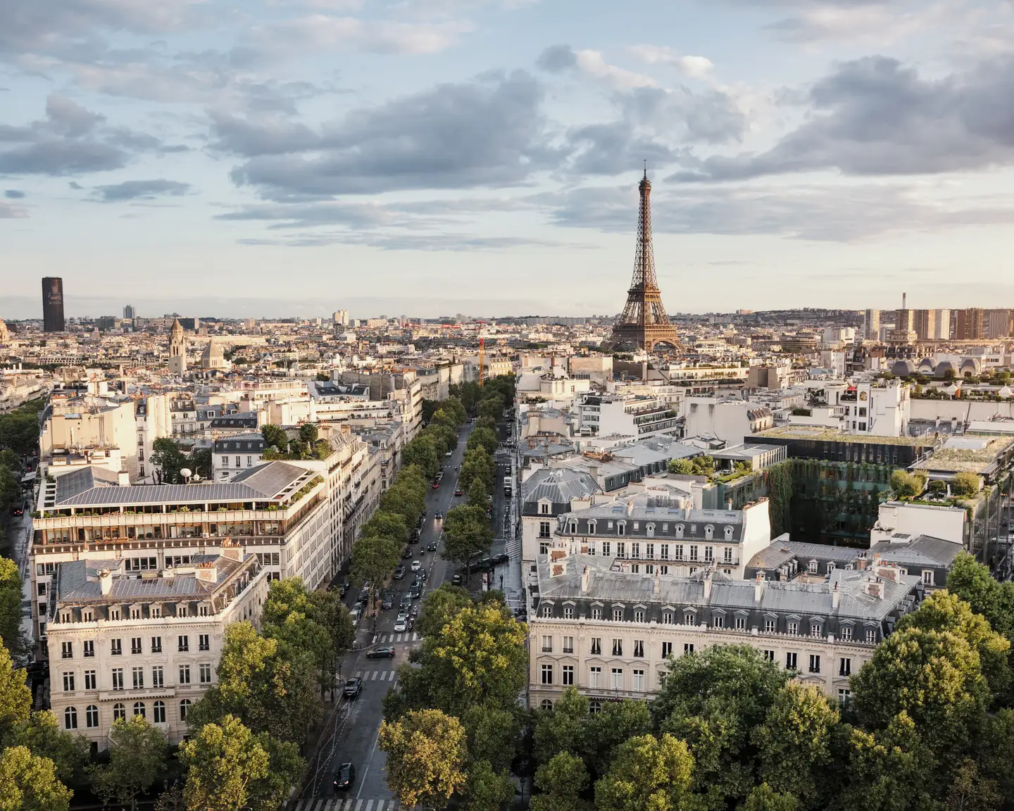 Panorama de Paris avec la Tour Eiffel, de larges avenues bordées d'arbres et une architecture parisienne classique sous un ciel partiellement nuageux.