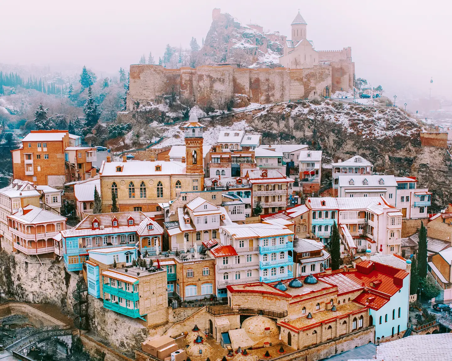 Vista invernal de un distrito histórico en Tiflis, Georgia. Se pueden ver coloridas casas georgianas con balcones de madera y fachadas de ladrillo cubiertas por una ligera capa de nieve. La fortaleza de Narikala, parcialmente cubierta por la nieve, se eleva al fondo, rodeada de árboles perennes envueltos en la niebla. 