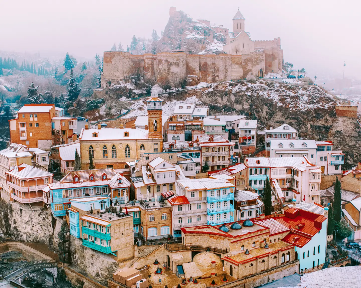 Une vue hivernale d'un quartier historique de Tbilissi, en Géorgie. On peut voir des maisons géorgiennes colorées aux balcons de bois et aux façades de briques recouvertes d’une légère couche de neige. La forteresse de Narikala, partiellement enneigée, se dresse à l'arrière-plan, entourée d'arbres brumeux à feuilles persistantes. 