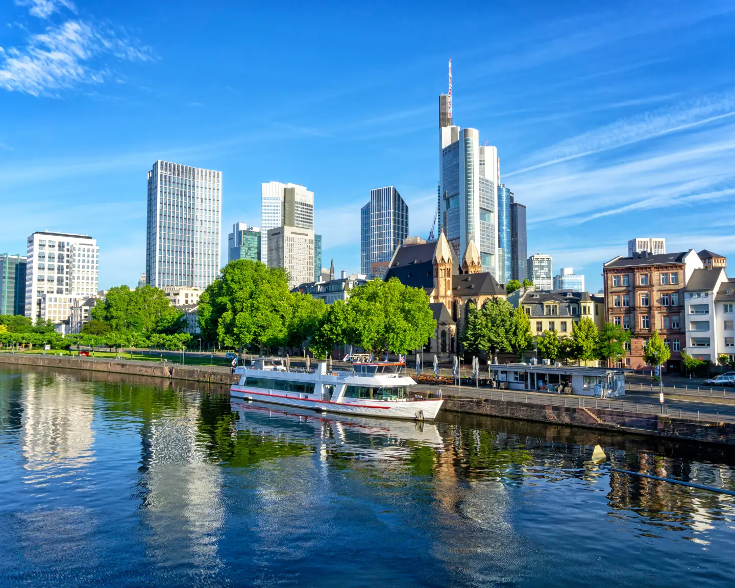 Skyline of Frankfurt am Main with modern skyscrapers, a river and an excursion boat under a clear blue sky.