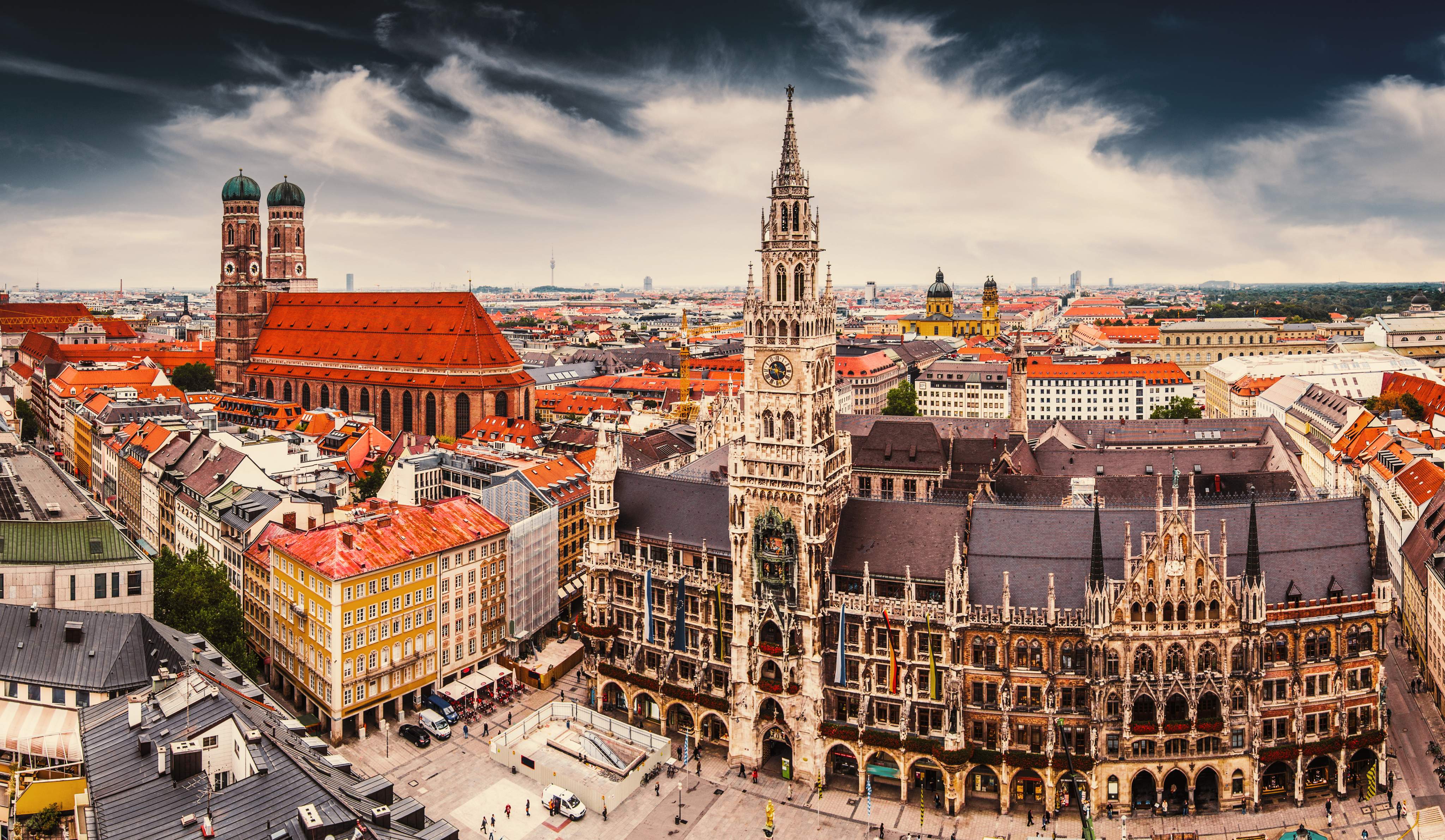 Panoramic view of the old town of Munich, Germany, with the New Town Hall and the Frauenkirche in the foreground under a dramatic sky.