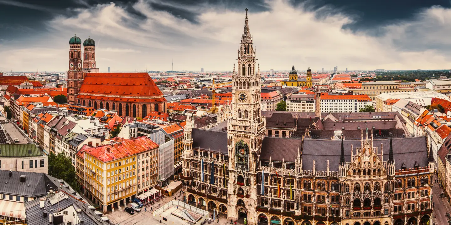Panoramic view of the old town of Munich, Germany, with the New Town Hall and the Frauenkirche in the foreground under a dramatic sky.