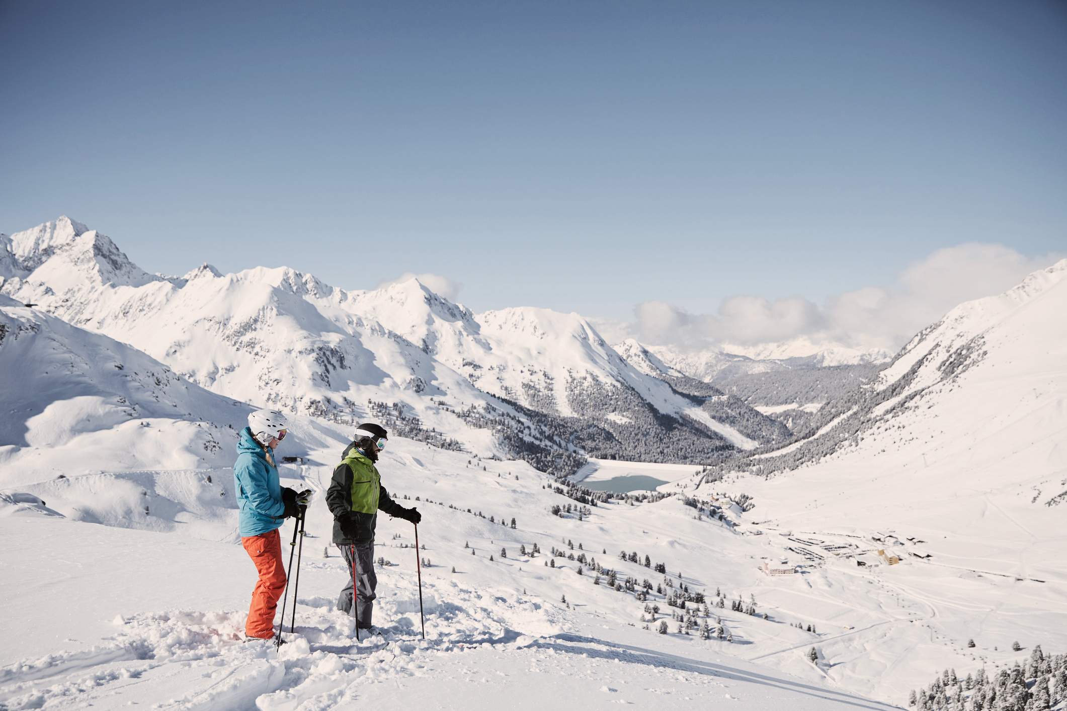Two skiers stand in the foreground looking down into the valley, with a snow-covered mountain landscape in the background.