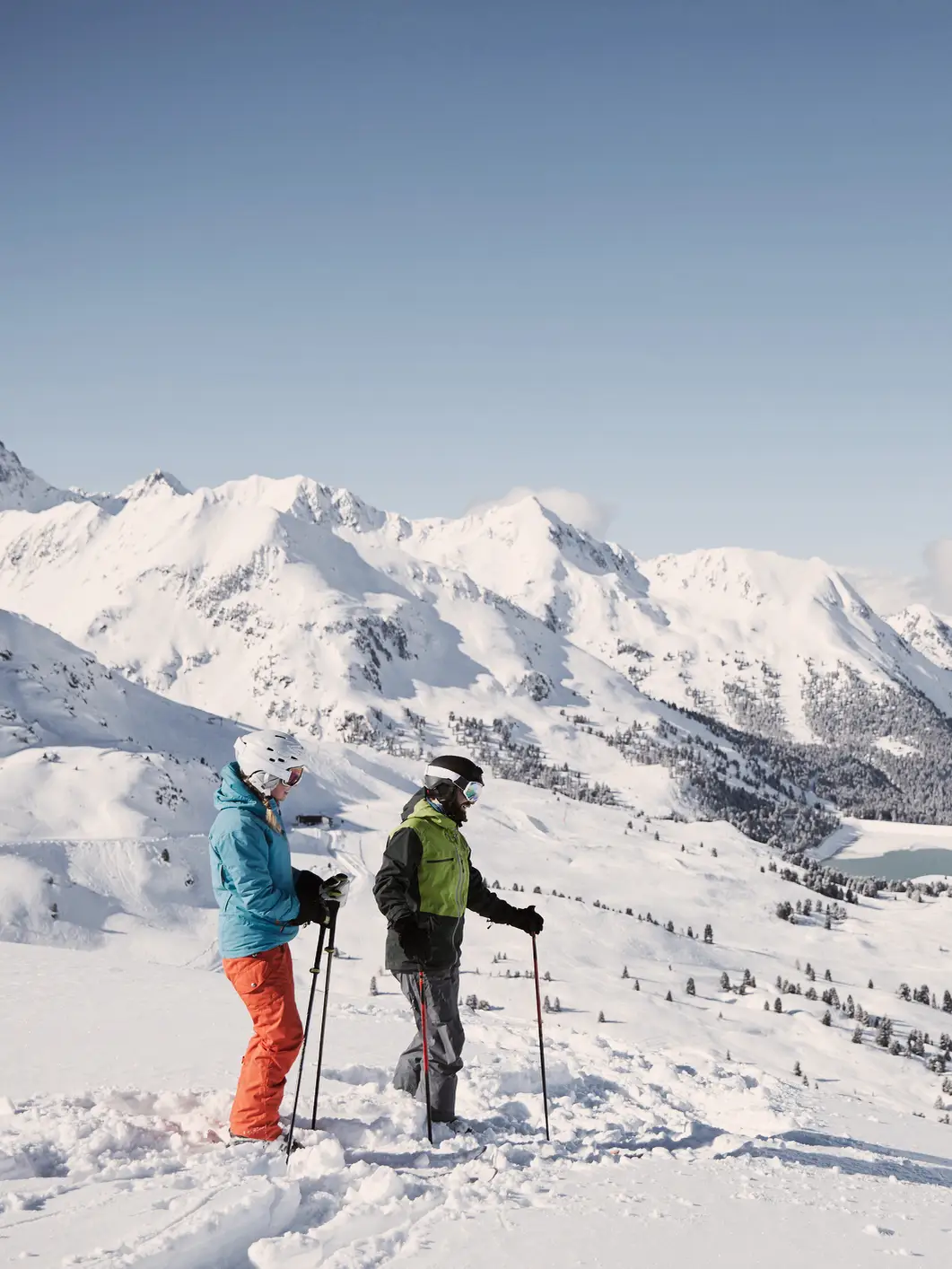 Two skiers stand in the foreground looking down into the valley, with a snow-covered mountain landscape in the background.