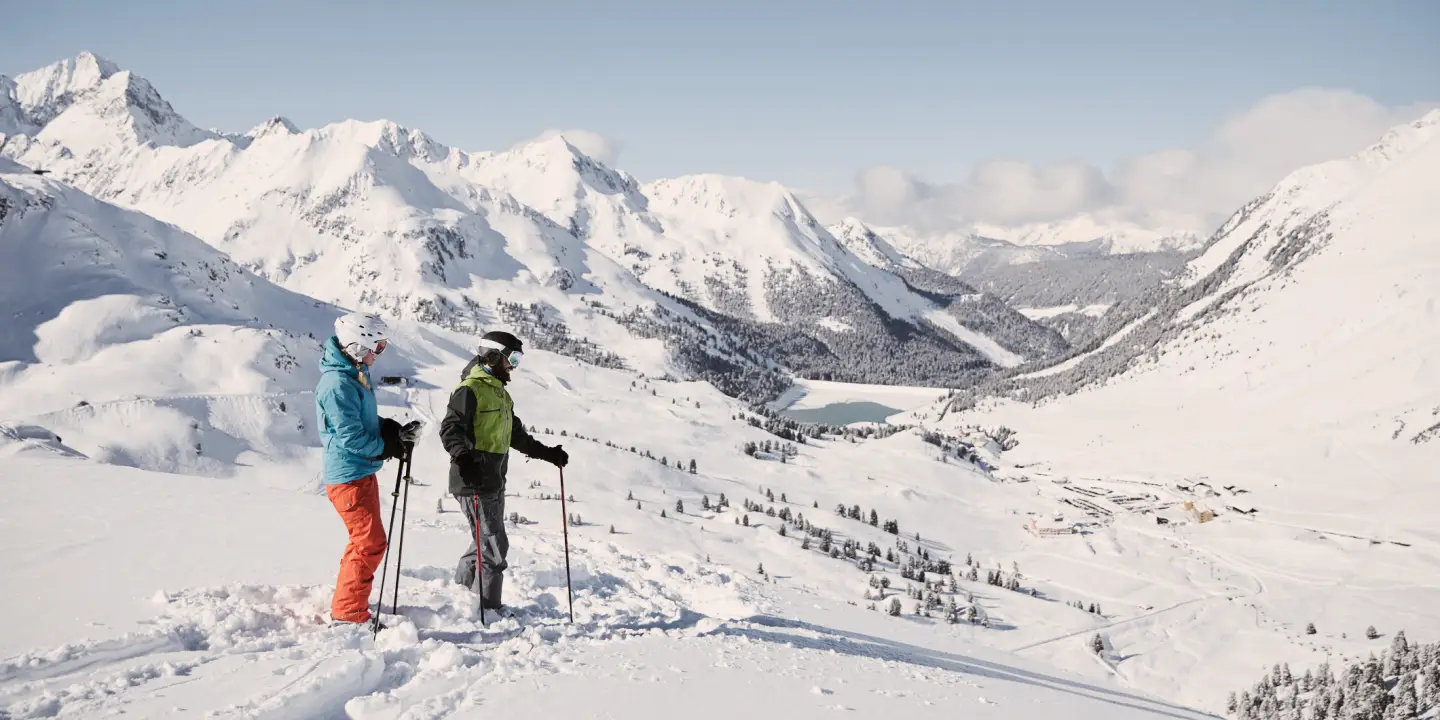 Two skiers stand in the foreground looking down into the valley, with a snow-covered mountain landscape in the background.