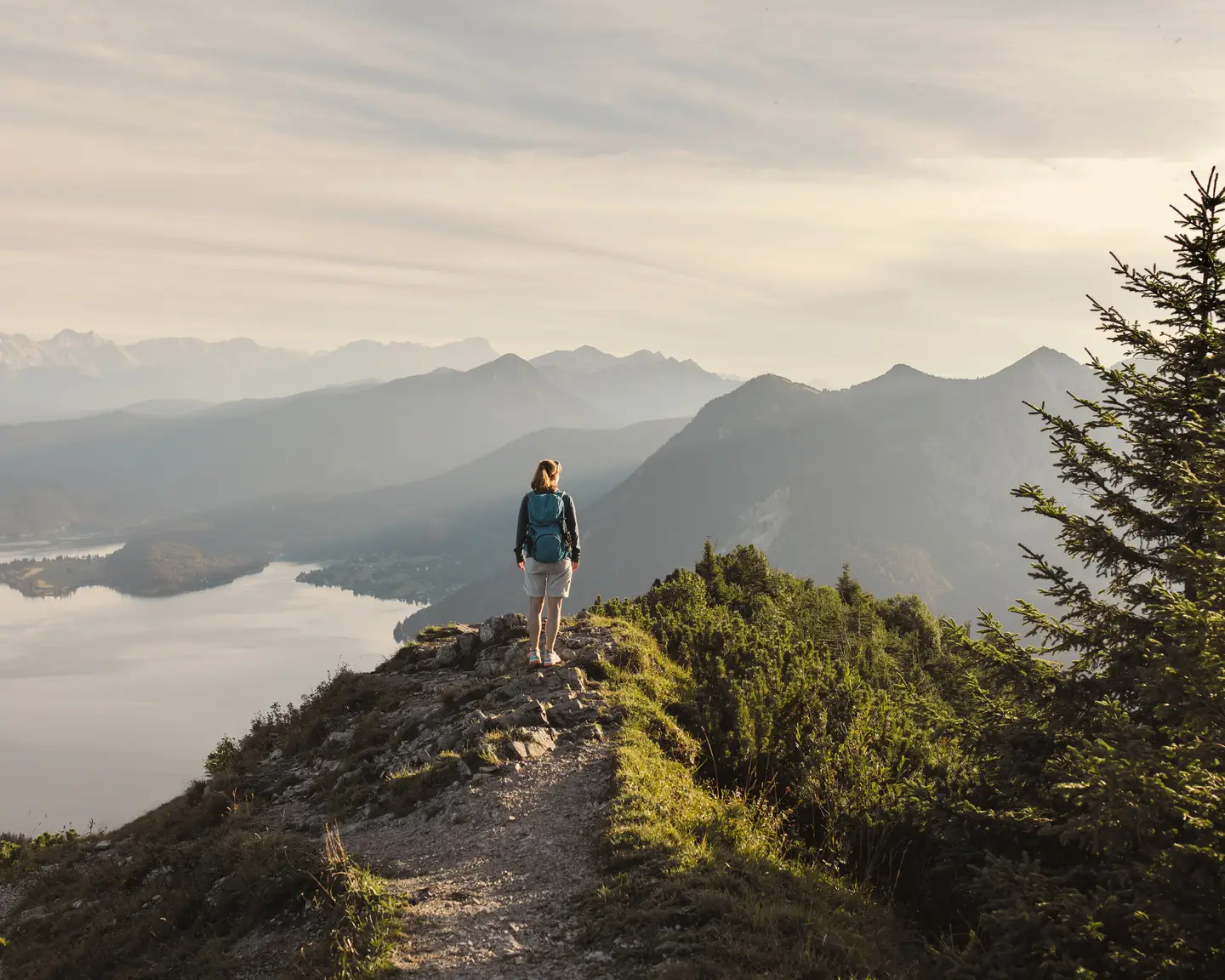 Mujer haciendo senderismo sola por una senda de montaña con vistas a un lago tranquilo y un panorama alpino.