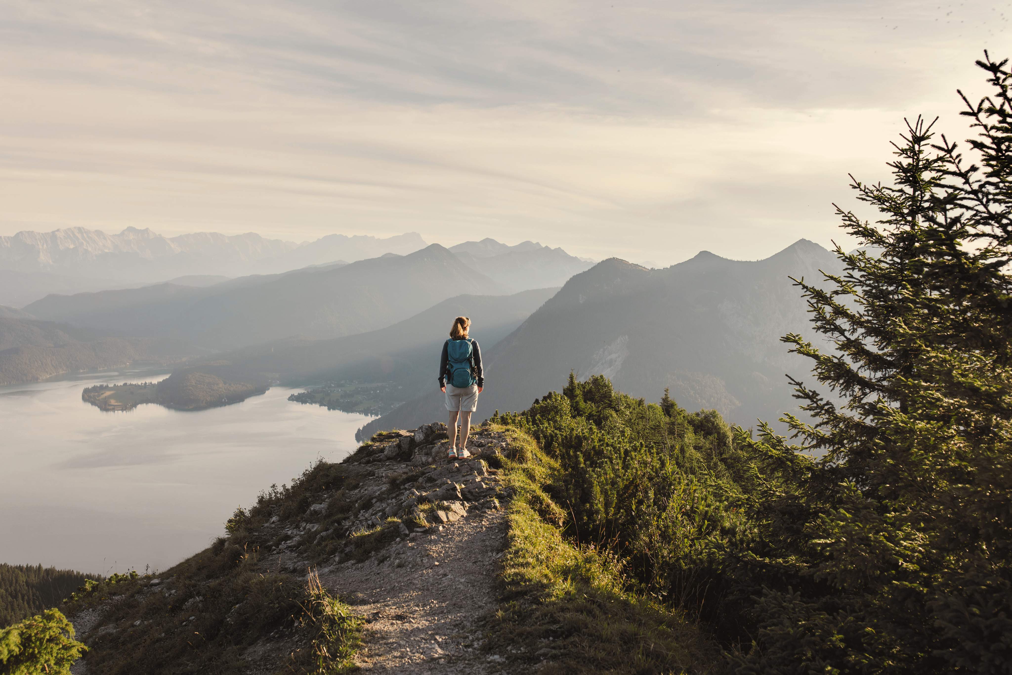 Woman hiking alone on a mountain trail with a view of a tranquil lake and an Alpine panorama.