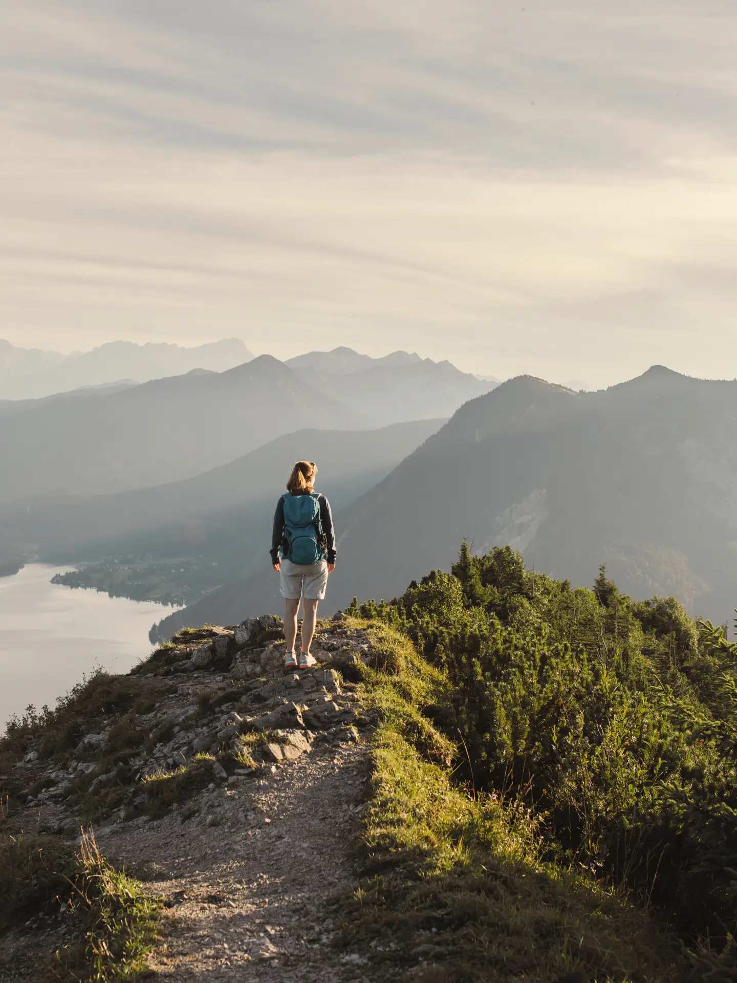 Woman hiking alone on a mountain trail with a view of a tranquil lake and an Alpine panorama.