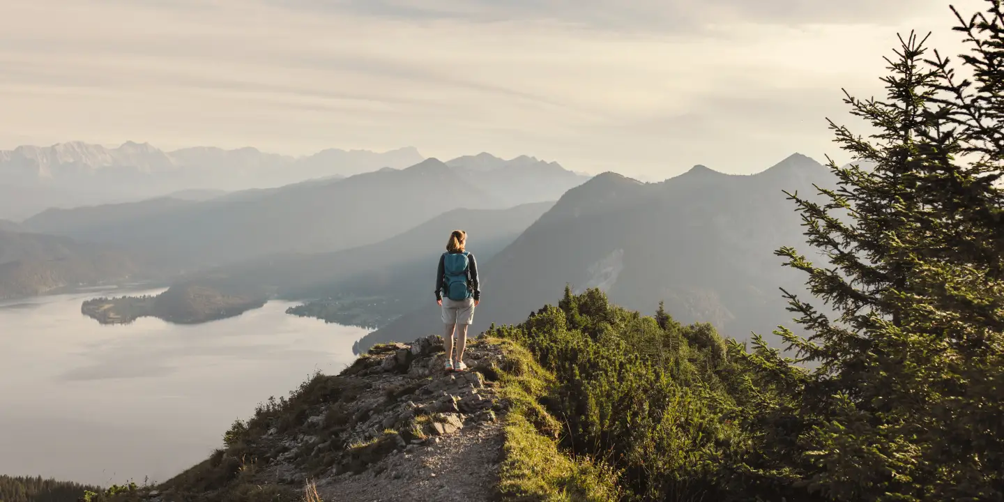 Woman hiking alone on a mountain trail with a view of a tranquil lake and an Alpine panorama.