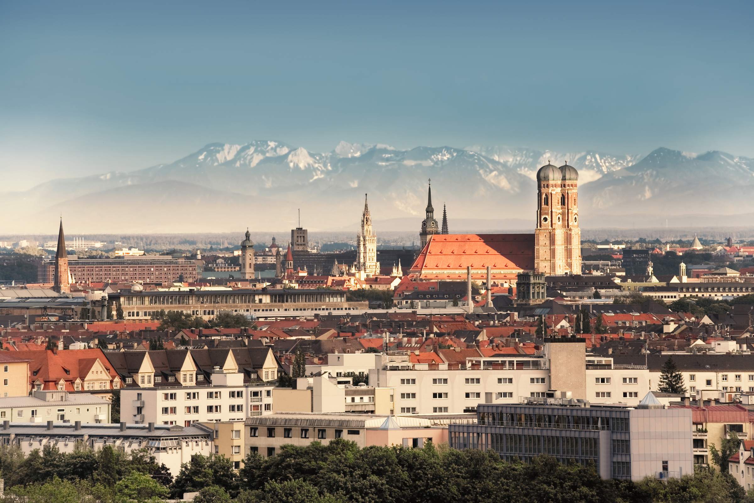 Panorama of Munich featuring the Frauenkirche and other historic buildings in the city center, with the Alps in the background under a clear sky.