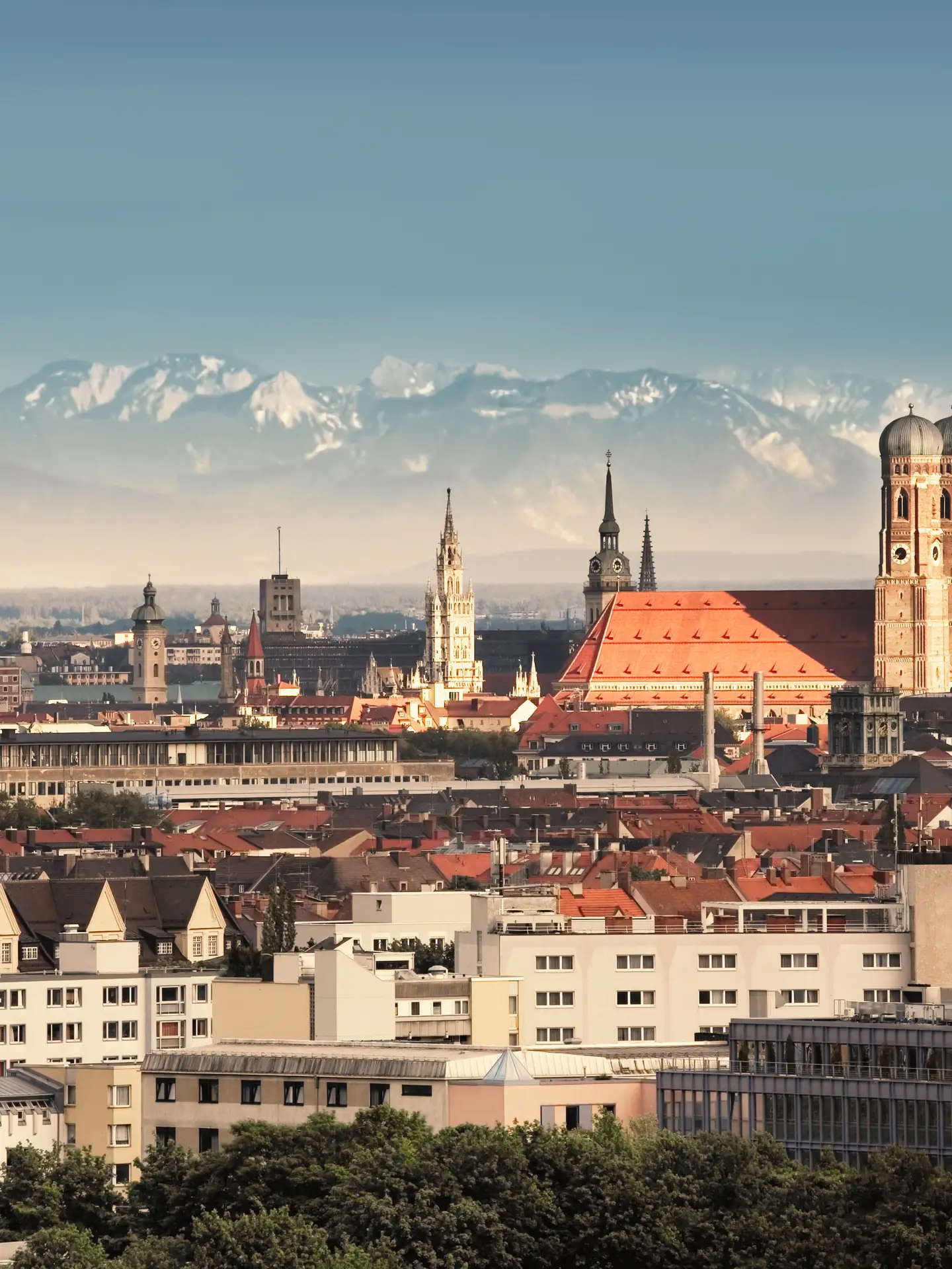 Panorama of Munich featuring the Frauenkirche and other historic buildings in the city center, with the Alps in the background under a clear sky.