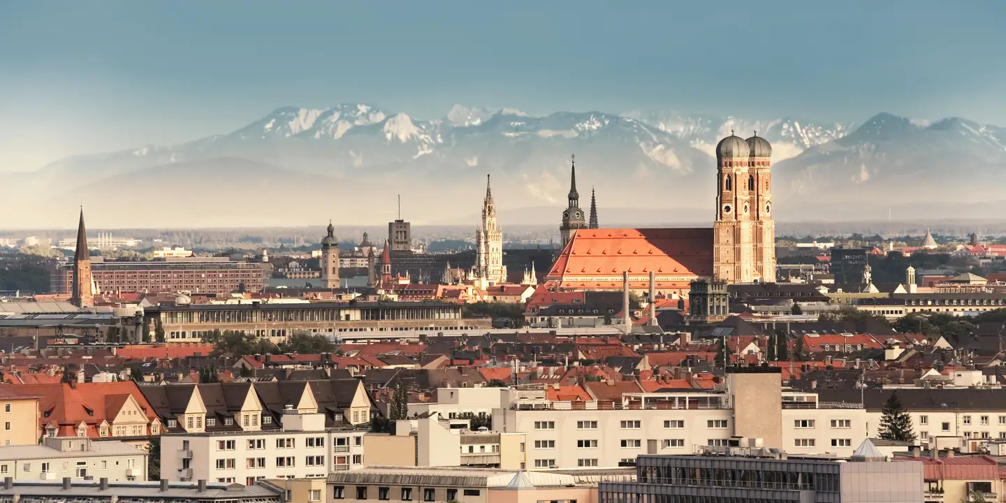 Panorama of Munich featuring the Frauenkirche and other historic buildings in the city center, with the Alps in the background under a clear sky.