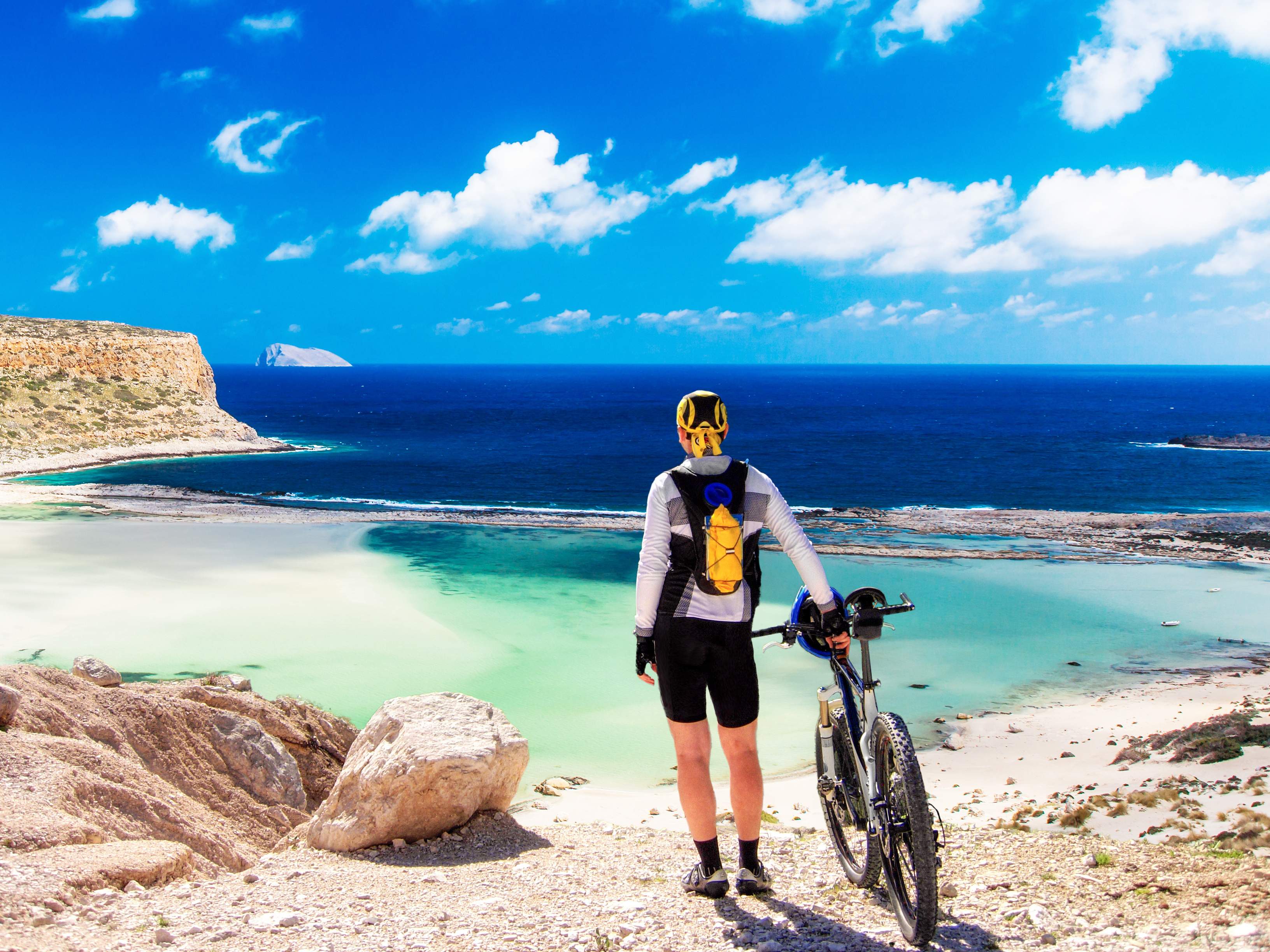 Ein Mountainbiker steht am Ufer der Balos Lagune auf Kreta, umgeben von kristallklarem Wasser und malerischen Felsenformationen.