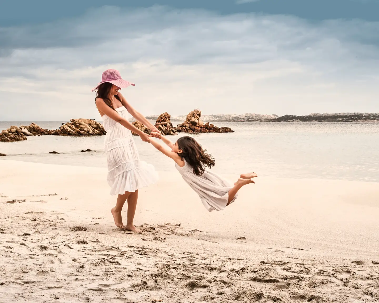 Una madre balancea a su hija en el aire en una playa. Ambas llevan un vestido blanco. Las rocas y el mar crean un pintoresco telón de fondo.