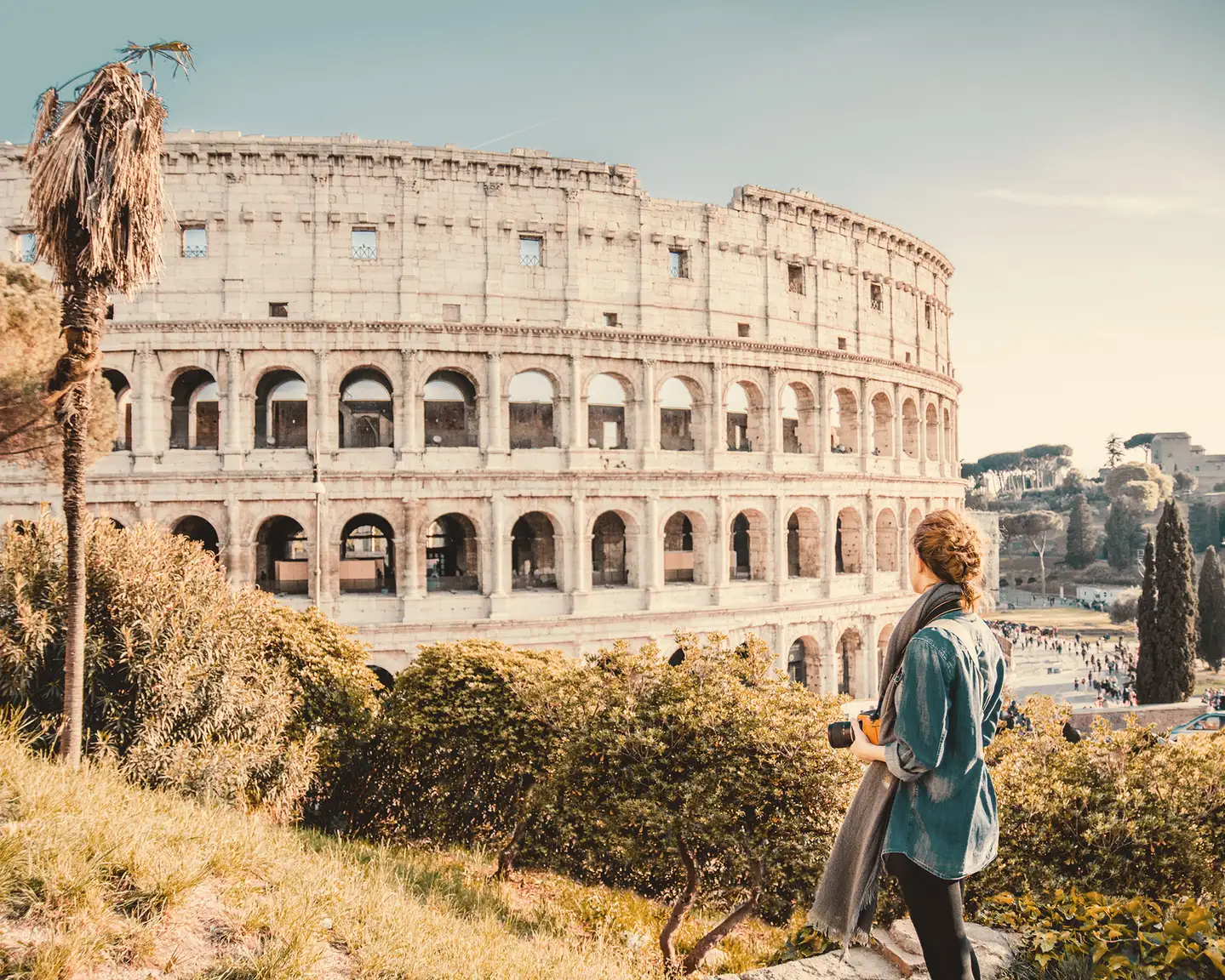 The Colosseum in Rome with a person in the foreground observing the historic structure with a camera in hand, surrounded by lush greenery.