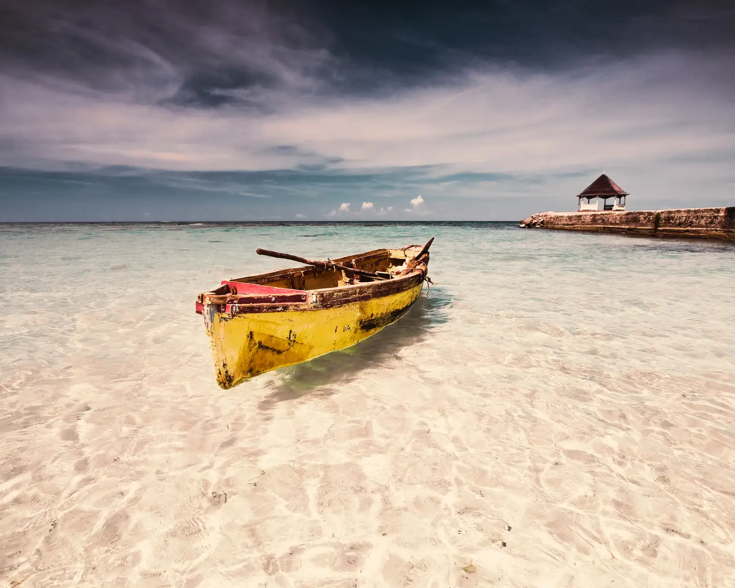 An old yellow boat on clear, calm water beside a beach in Jamaica, with a small hut on a jetty in the background.
