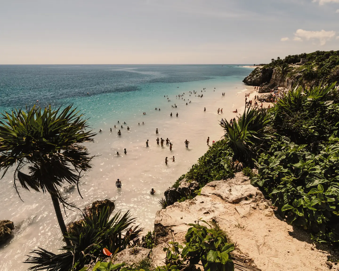 Blick von einer Felsklippe mit vielen Büschen auf einen wunderschönen Strand, an dem viele Menschen die Sonne im Wasser genießen.