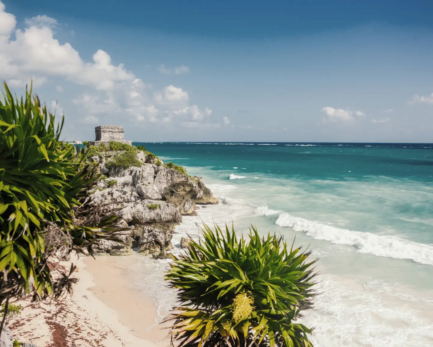 Pohled na pobřeží u Tulumu v Cancúnu s mayskými ruinami na útesu, obklopenými tropickými rostlinami a tyrkysovým mořem.