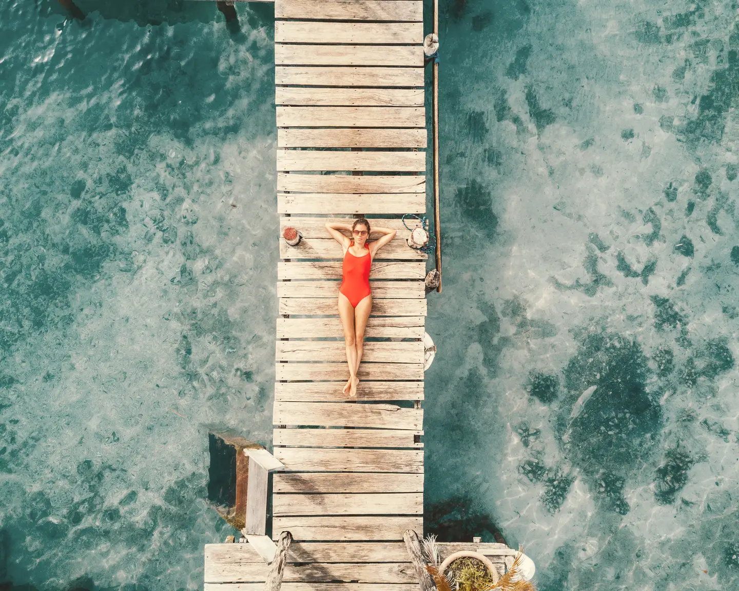Vista aérea de una mujer en un traje de baño rojo relajándose en un puente de madera sobre agua transparente y turquesa.