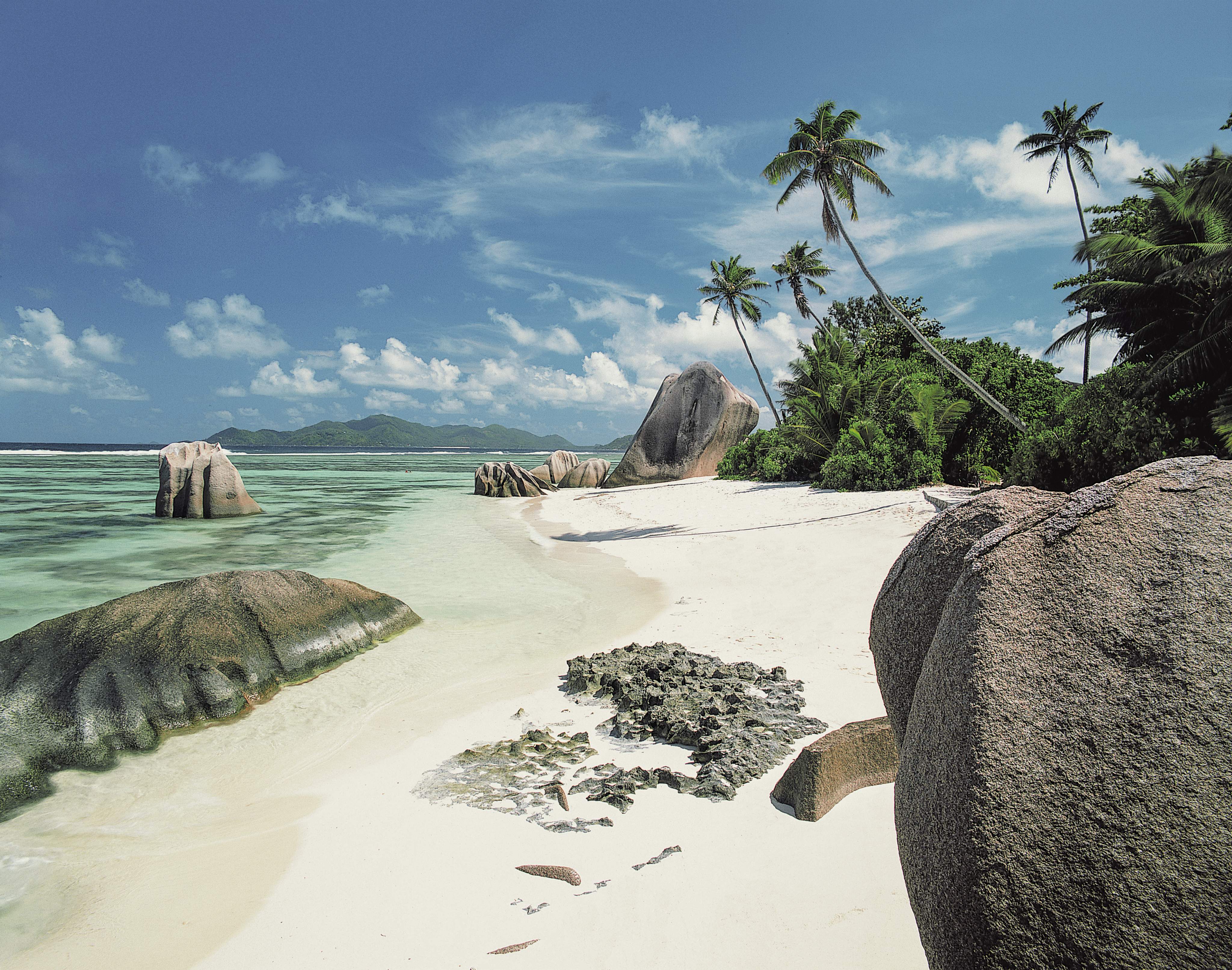 La plage de l'Anse Source d'Argent à La Digue, aux Seychelles, avec du sable blanc, de grands rochers de granit, des palmiers et une eau claire et turquoise sous un ciel bleu.