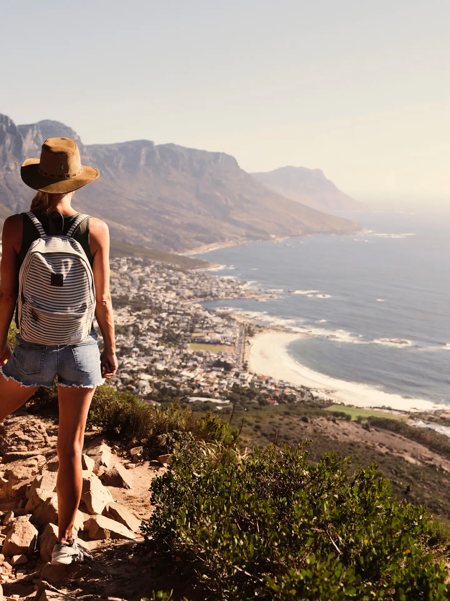 Mujer con mochila y sombrero de pie sobre el pico Cabeza de León, contemplando la costa de Ciudad del Cabo, Sudáfrica.