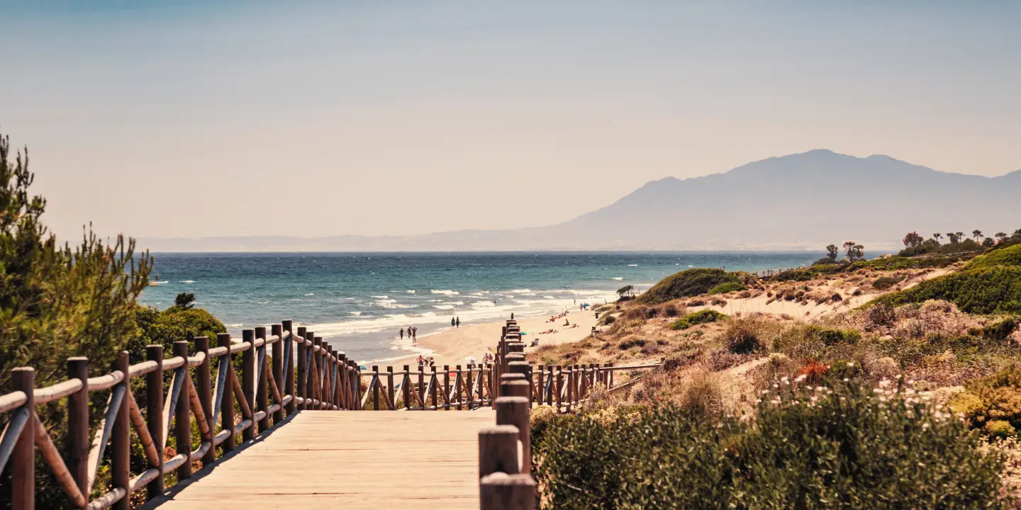 Une plage à Málaga, Espagne, avec une passerelle en bois menant à la plage de sable et la mer et les montagnes en arrière-plan, sous un ciel bleu clair.