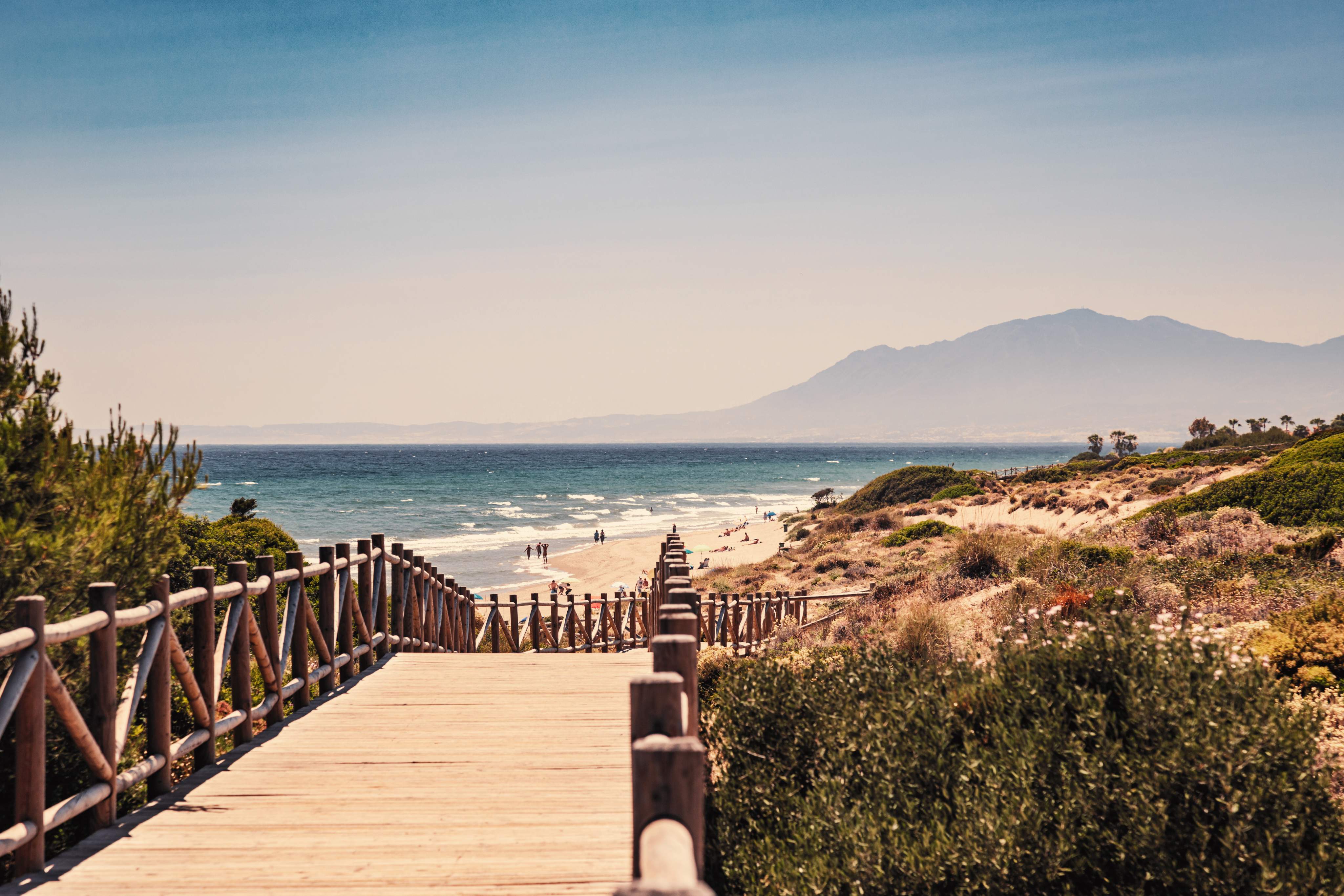 A beach in Málaga, Spain, with a wooden walkway leading to the sandy beach and the sea and mountains in the background, beneath a clear blue sky.