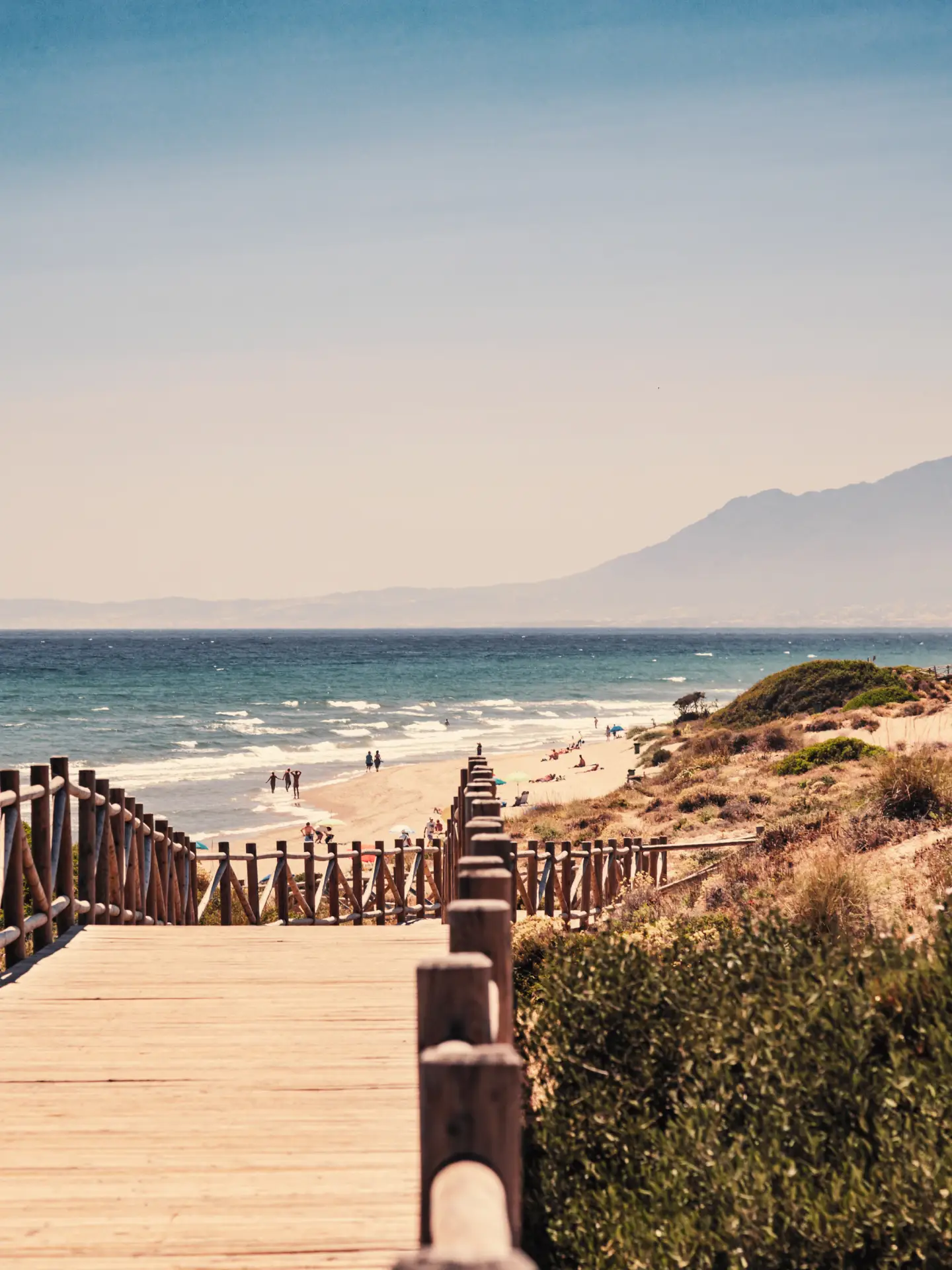 A beach in Málaga, Spain, with a wooden walkway leading to the sandy beach and the sea and mountains in the background, beneath a clear blue sky.