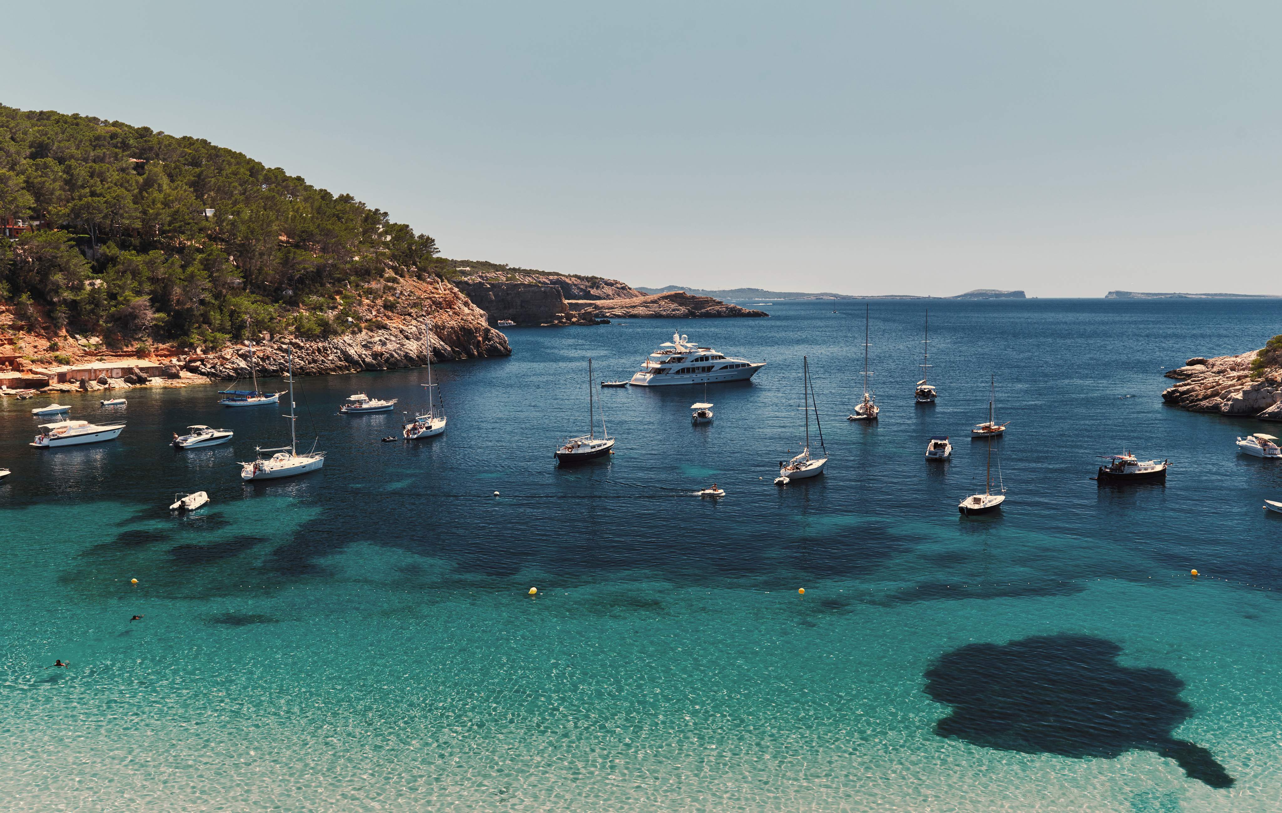Blick auf eine türkisfarbene Bucht auf Ibiza mit Segelbooten, Yachten und der felsigen Mittelmeerküste unter blauem Himmel.