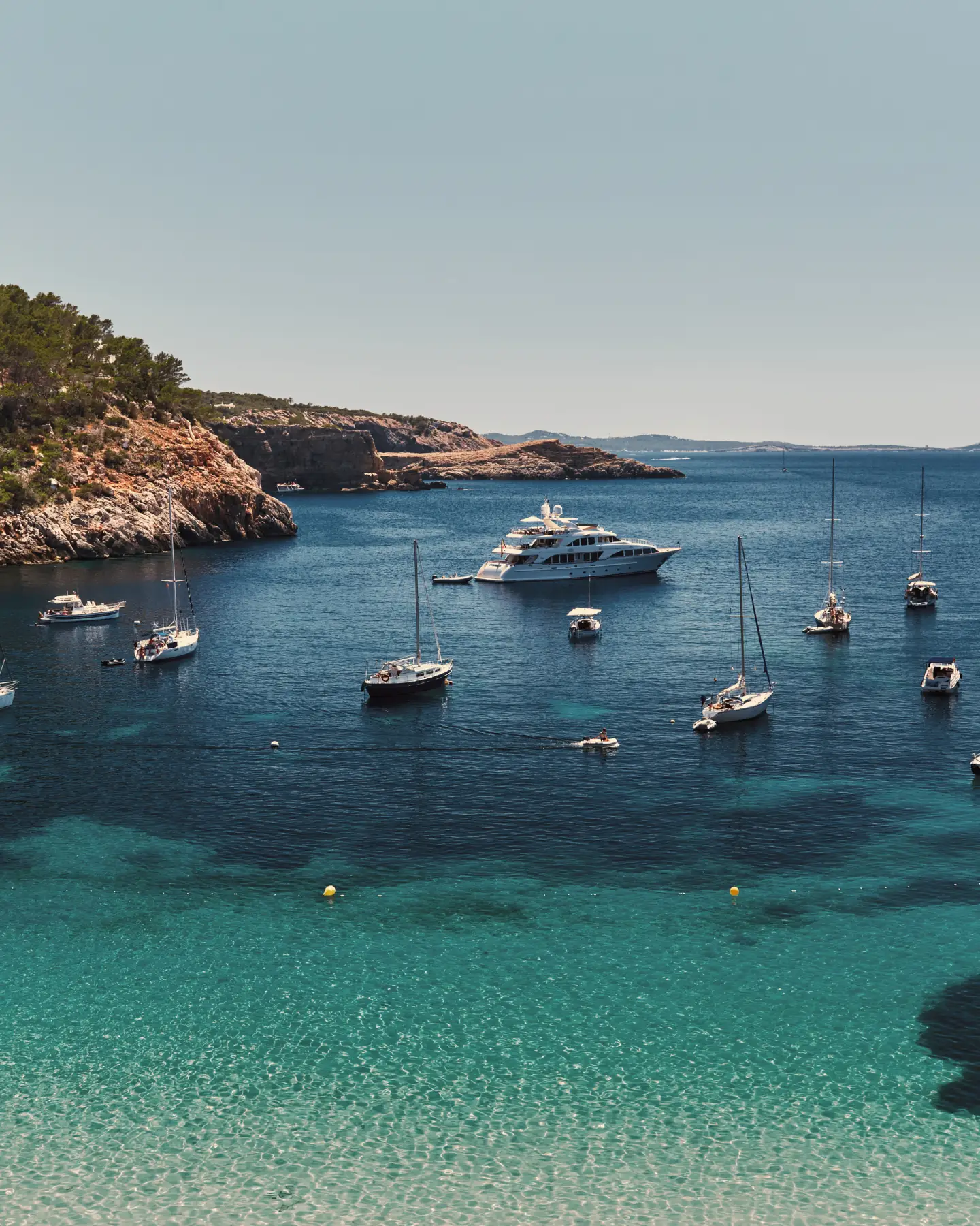 Blick auf eine türkisfarbene Bucht auf Ibiza mit Segelbooten, Yachten und der felsigen Mittelmeerküste unter blauem Himmel.