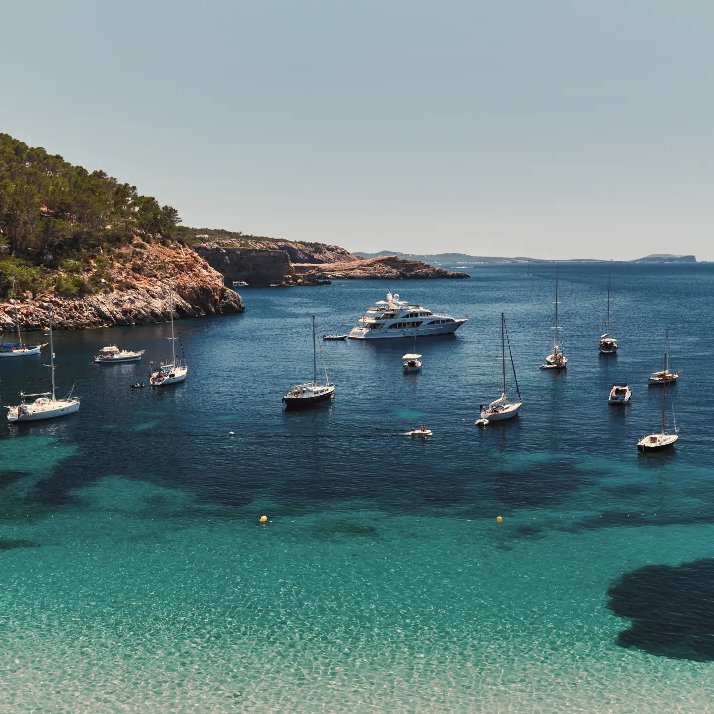 Blick auf eine türkisfarbene Bucht auf Ibiza mit Segelbooten, Yachten und der felsigen Mittelmeerküste unter blauem Himmel.