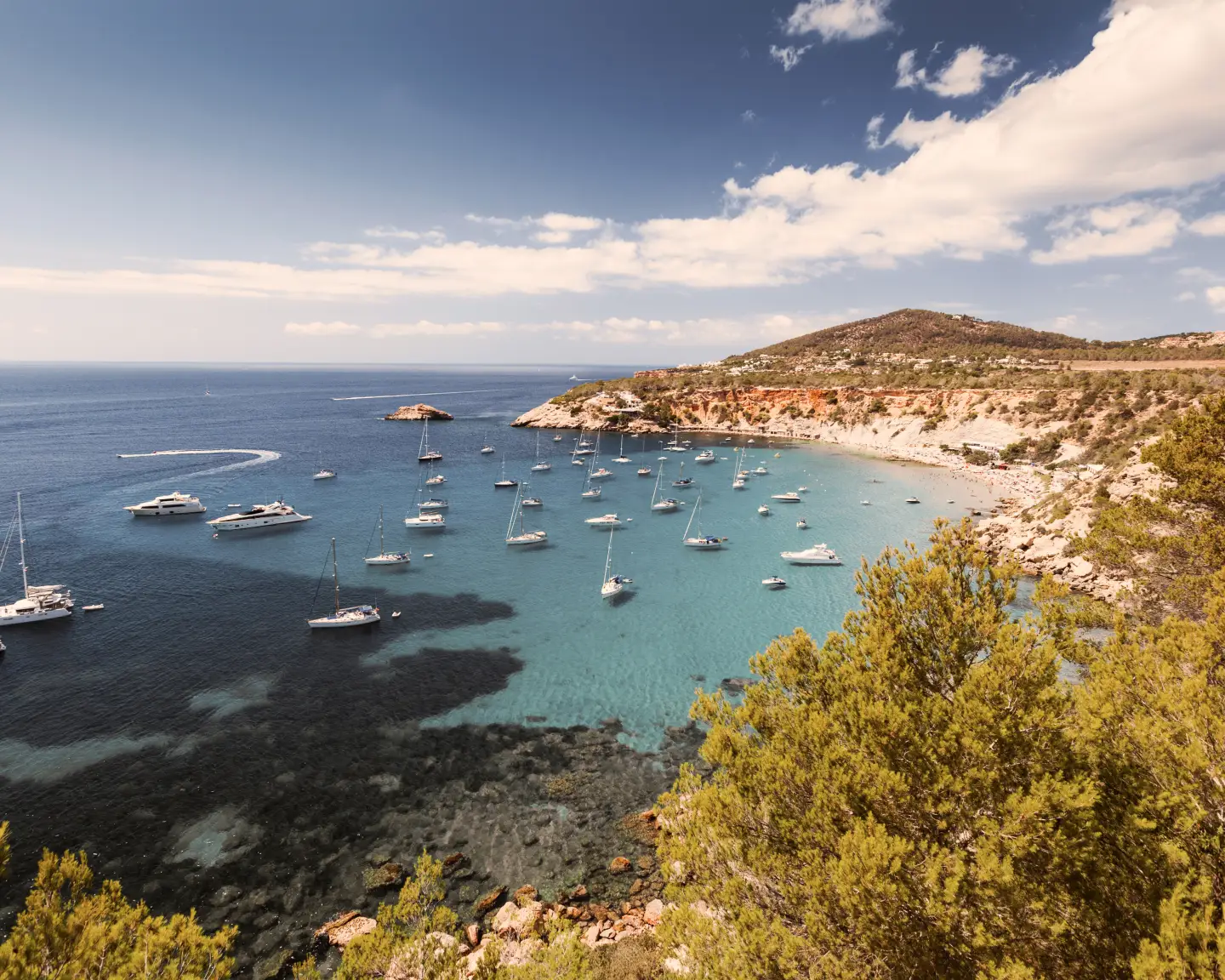 Küstenbucht auf Ibiza mit türkisblauem Wasser, Segelbooten und grüner Felslandschaft.