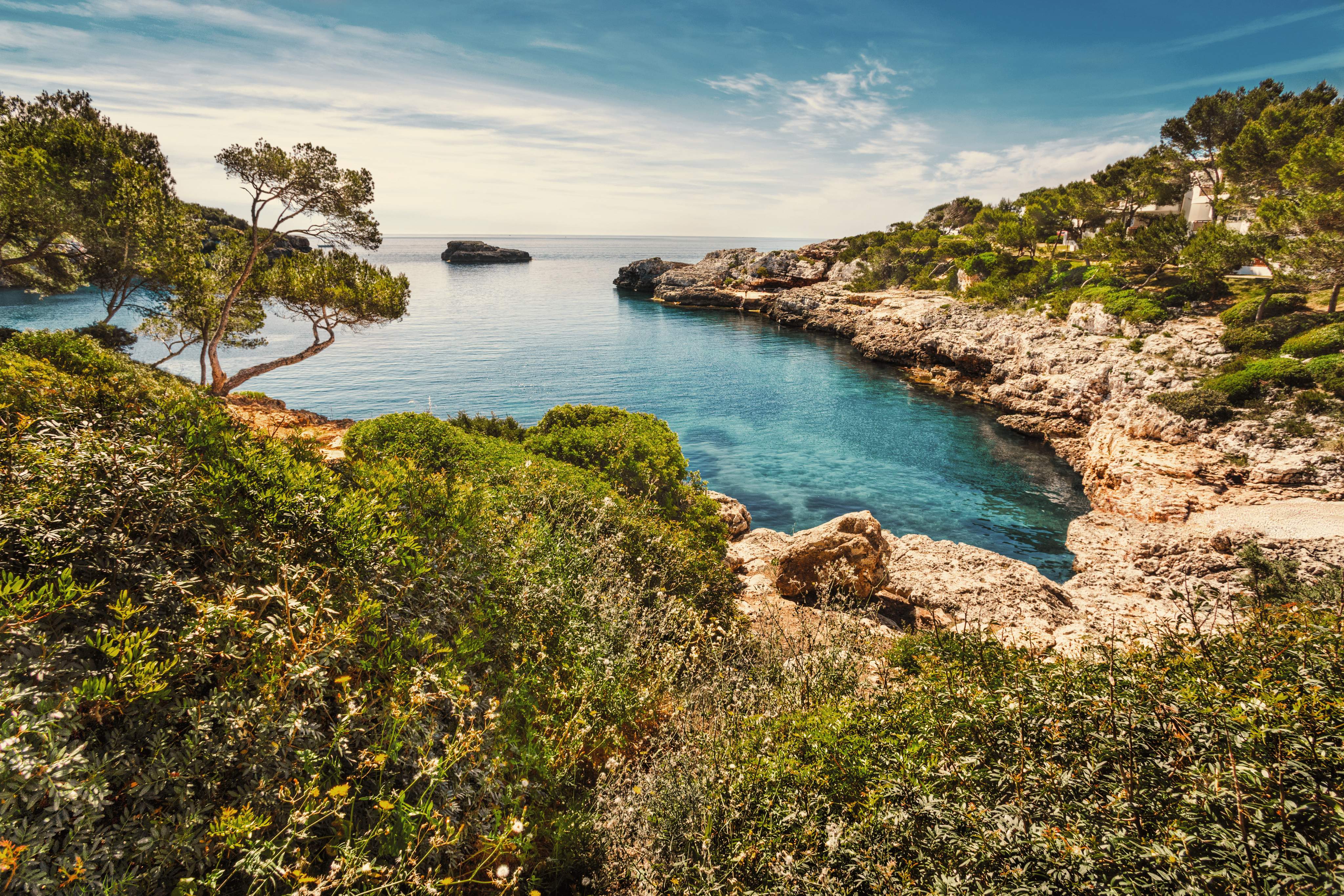 Pittoreske baai in Mallorca, Spanje, met turquoiseblauw water, ruige rotsen en groene mediterrane vegetatie onder een blauwe hemel.