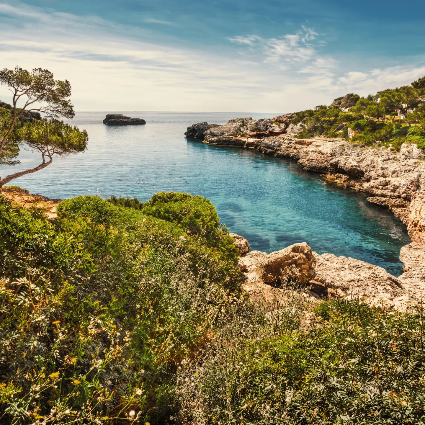 Pittoreske baai in Mallorca, Spanje, met turquoiseblauw water, ruige rotsen en groene mediterrane vegetatie onder een blauwe hemel.