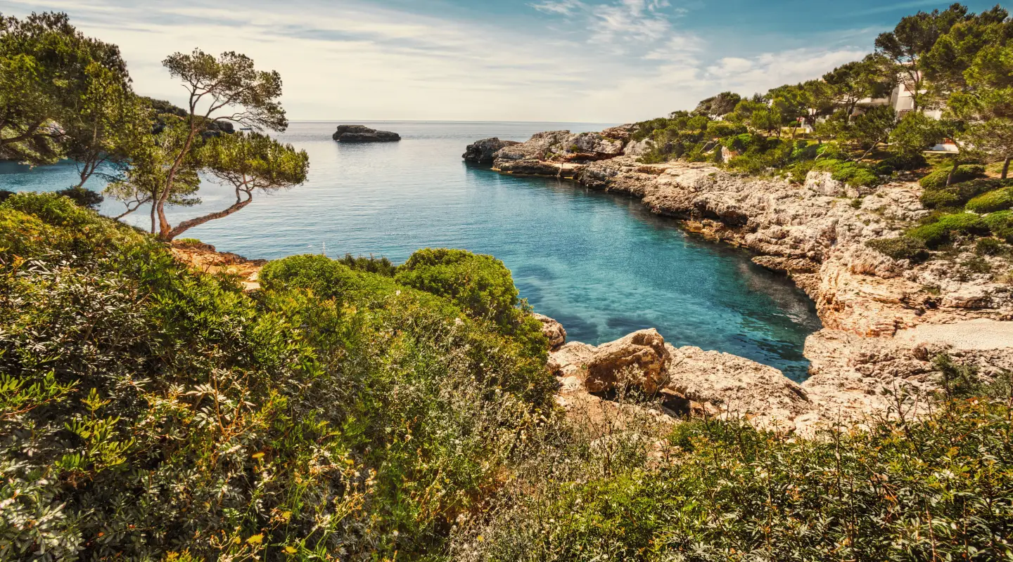 Pittoreske baai in Mallorca, Spanje, met turquoiseblauw water, ruige rotsen en groene mediterrane vegetatie onder een blauwe hemel.