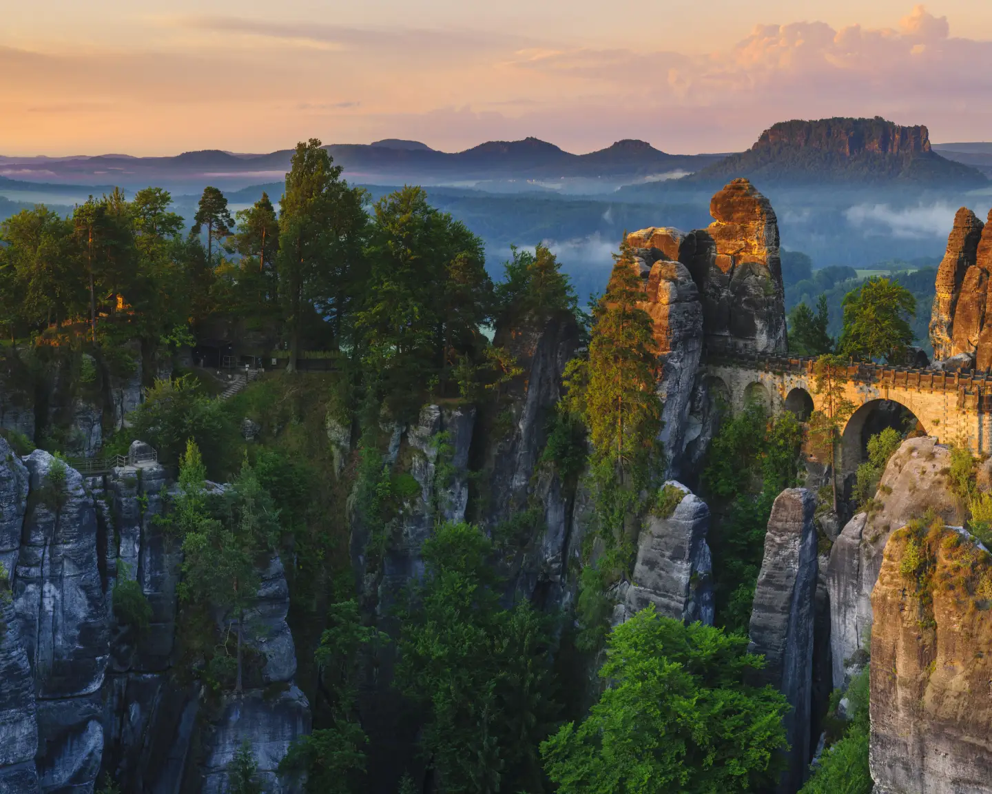 Panoramablick auf die Basteibrücke im Nationalpark Sächsische Schweiz mit markanten Sandsteinfelsen, dichtem Wald und Abendstimmung.