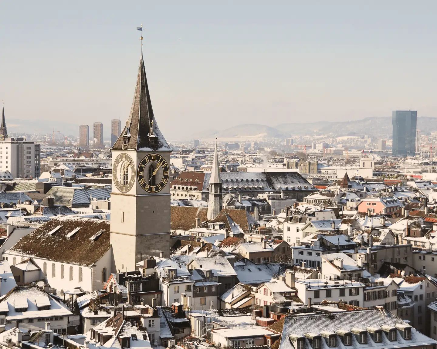 Vue de Zurich en hiver avec l'église Saint-Pierre et sa grande horloge, les toits enneigés de la vieille ville et les gratte-ciel modernes en arrière-plan.