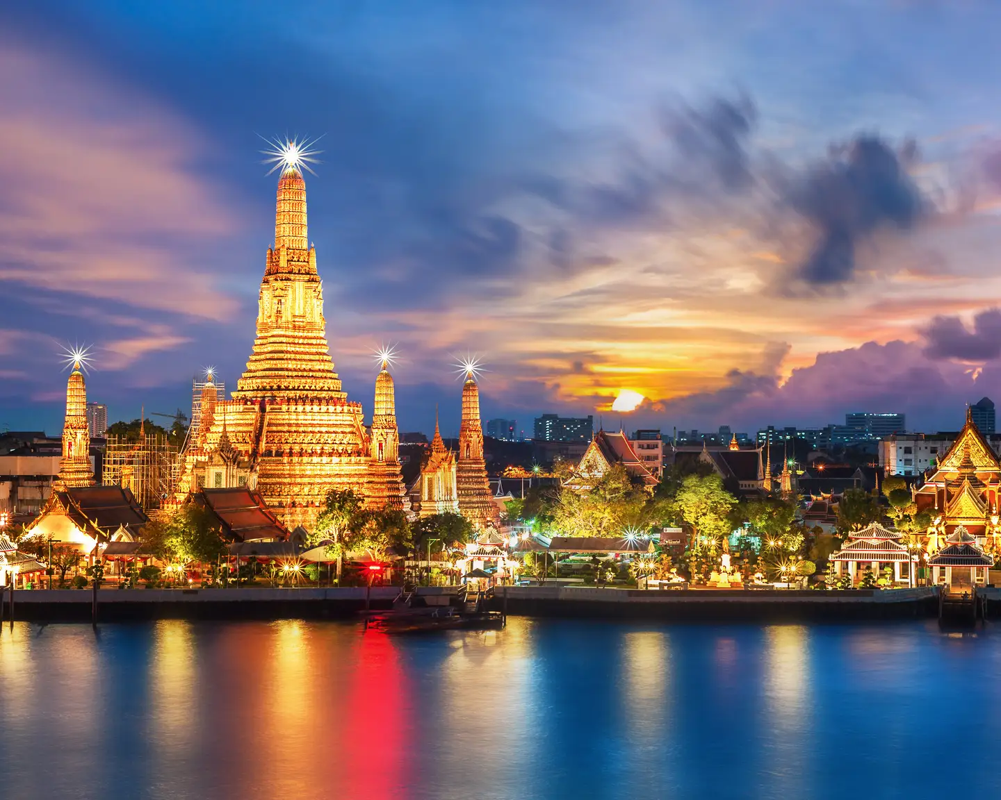 El templo Wat Arun iluminado en Bangkok al atardecer, con cielos coloridos y reflejos en el agua del río Chao Phraya.