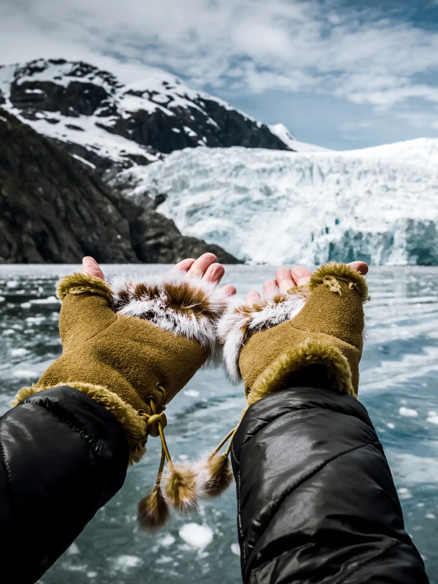 Persona con guantes gruesos y chaqueta de invierno extendiendo las manos hacia un glaciar rodeado de un lago y enmarcado entre montañas nevadas.