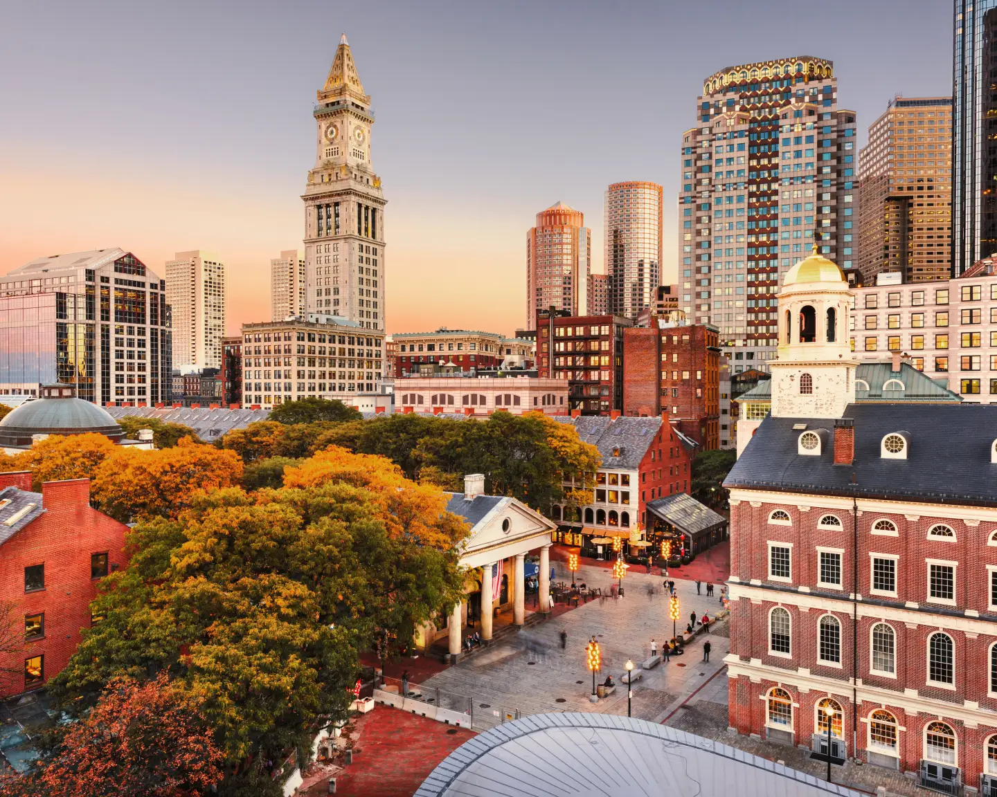 Perfil urbano de Boston al atardecer con la histórica Custom House Tower y edificios al fondo, y una plaza con árboles otoñales en primer plano.