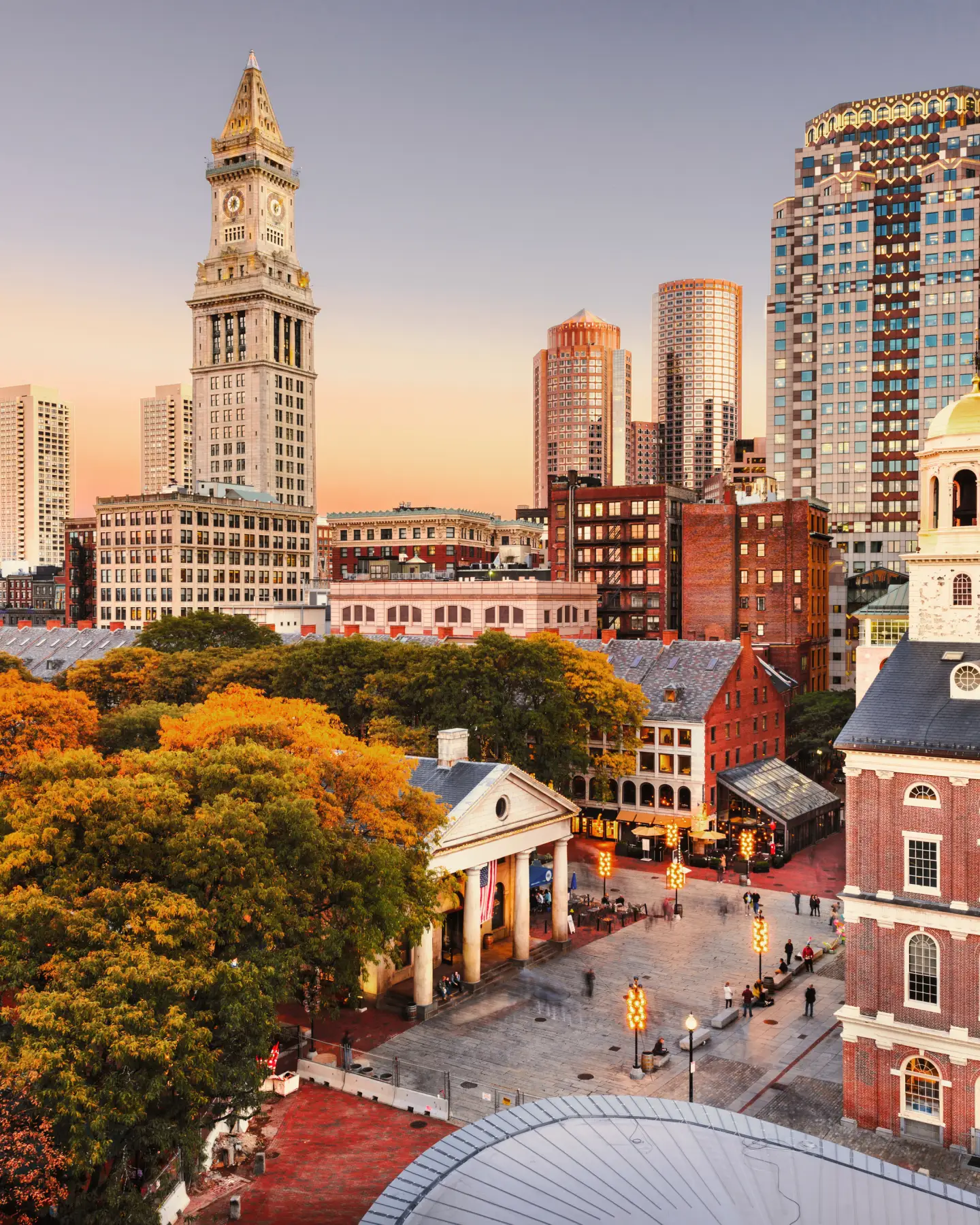 Panorama de Boston au coucher du soleil, avec la tour historique Custom House et des bâtiments en arrière-plan, et une place avec des arbres aux couleurs automnales au premier plan.