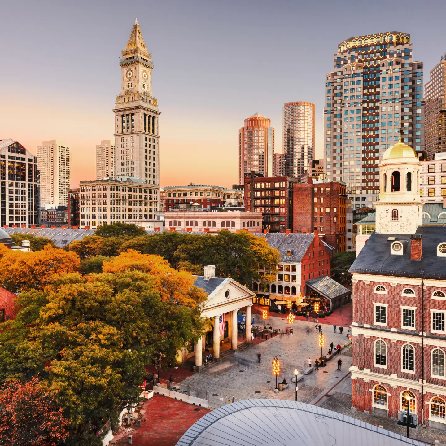 Städtische Skyline von Boston bei Sonnenuntergang mit dem historischen Custom House Tower und Gebäuden im Hintergrund sowie einem Platz mit Herbstbäumen im Vordergrund.