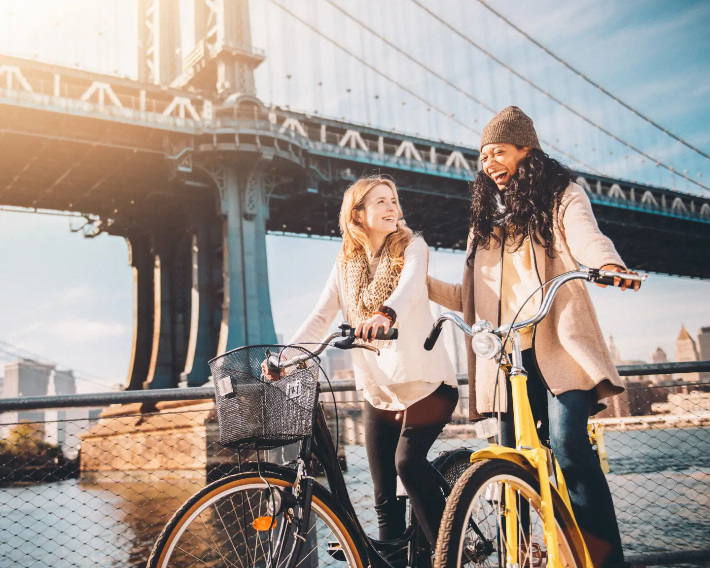 Unas amigas se ríen en bicicleta bajo el puente de Manhattan con una vista de Brooklyn y el perfil urbano de fondo.