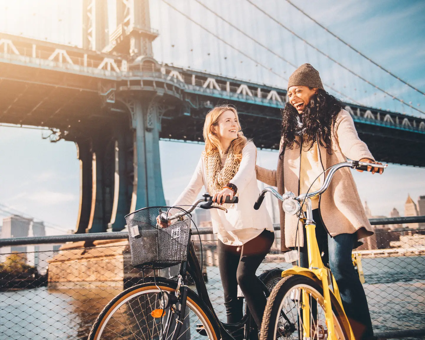 Unas amigas se ríen en bicicleta bajo el puente de Manhattan con una vista de Brooklyn y el horizonte de fondo.