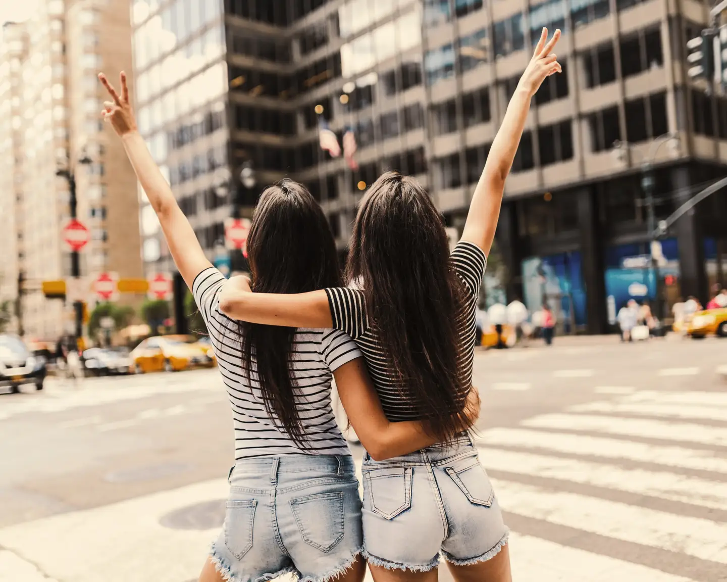 Dos mujeres de cabello oscuro están de pie en medio de una intersección en Nueva York; se las puede ver desde atrás con los brazos alrededor de los hombros de la otra, cada una sosteniendo su mano libre en el aire con el signo de la paz.