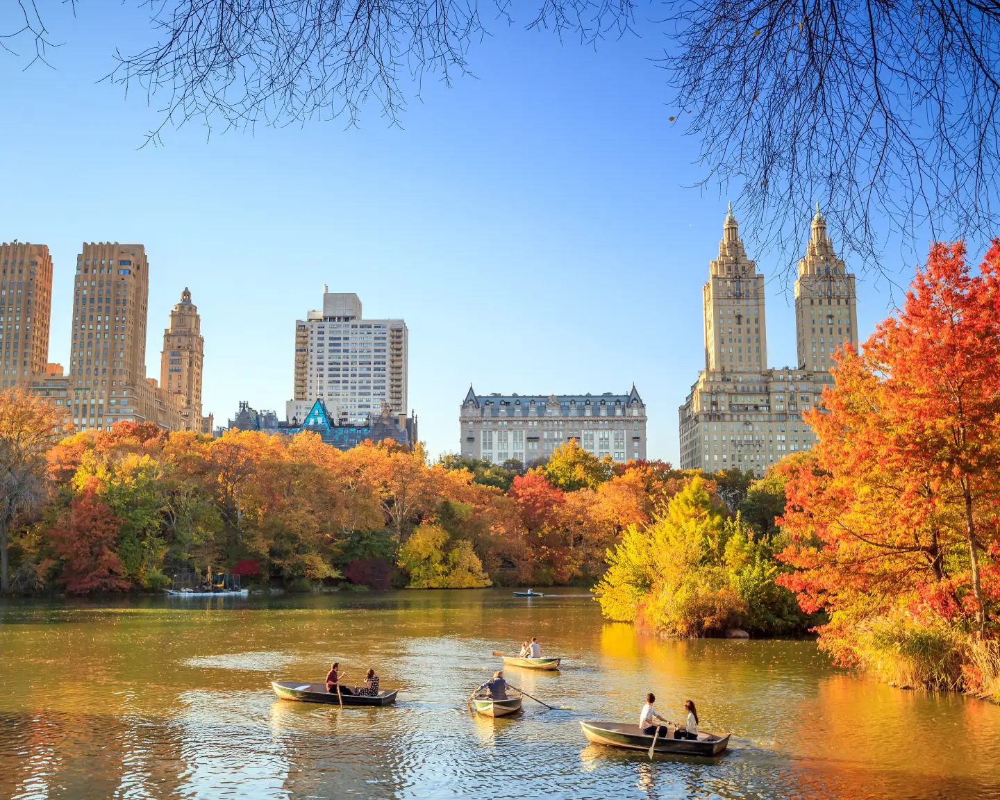 Autumn scene in Central Park, New York City, with people in rowing boats on the lake, surrounded by colourful autumn trees and the urban skyline in the background.