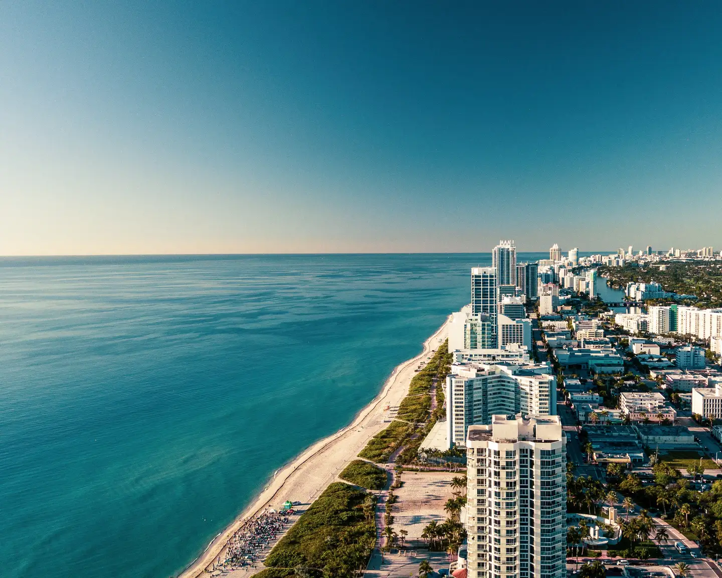 Aerial view of Miami Beach with tall buildings along the coast, a fine sandy beach, and the vast blue ocean under a clear sky.