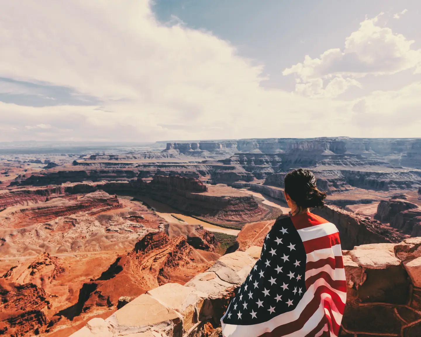Eine Frau mit einer amerikanischen Flagge um die Schultern blickt auf den Grand Canyon, unter einem weiten, sonnigen Himmel.